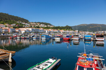 A serene marina of Muros in Spain with various boats docked in calm waters, with quaint hillside houses and clear blue sky in the background - obrazy, fototapety, plakaty
