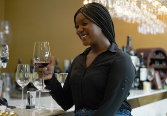 Black female tourist sits holding a glass of red mattos wine, happily tasting wine at the counter...
