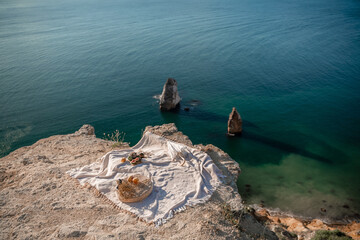 A white blanket is spread out on a beach near the water. The blanket is on a rock and there are some oranges and apples on it. The scene is peaceful and relaxing, with the ocean in the background.