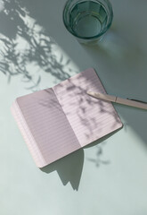 A Notebook with a pen on the blue table with leaves shadows. Copy space