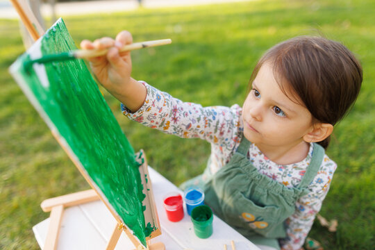 Little girl is painting a picture in the park on the green grass. Child development concept.