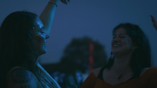 Two female friends wearing glitter holding drinks having fun dancing at outdoor summer music festival at night - shot in slow motion 