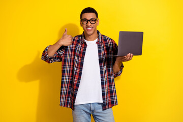 Portrait of nice young man hold laptop show thumb up wear shirt isolated on yellow color background