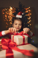Fototapeta na wymiar Happy 6 years old girl in Santa Claus costume wraps gifts boxes with red ribbons and festive lights on background