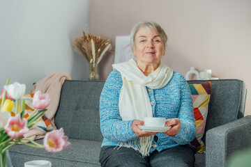Smiling mature woman holding cup of tea, relaxing at home, positive senior female sitting on couch in modern living room looking satisfied feels good, older generation happy retired time enjoy life.