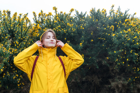 Smiling young woman in a yellow raincoat and hood with closed eyes enjoying the moment on walk with blooming bushes. Travel concept. Hiking in any weather. Girl on a journey. personal fulfillment