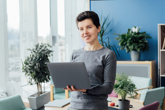 Portrait of Smiling Neutral gender middle aged woman in casual outfit working on laptop while standing in modern office, meeting room, conference space. Business process organization. Comfy workplace.