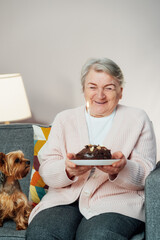 Cheerful smiling mature aged senior woman holds birthday cake and blowing a candle, celebrates a birthday at home with dog pet and laughs, feeling happy. Enjoying retirement lifestyle. Vertical card