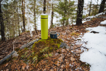 A light green thermos stands on a stone in the forest next to a metal mug, a concept of tourist...