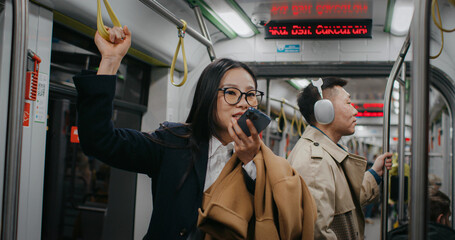 Serious Asian businesswoman holding on to safety handle with one arm. Recording voice message to someone with instructions. People in background standing still and listening to music in headphones.