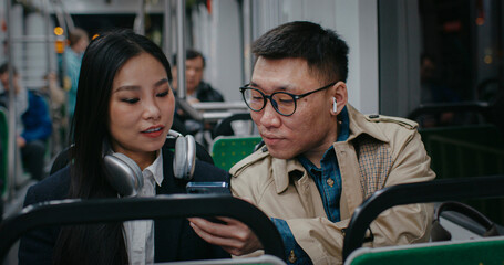Cute Chinese couple sitting next to each other on fast moving train. Man showing something to...
