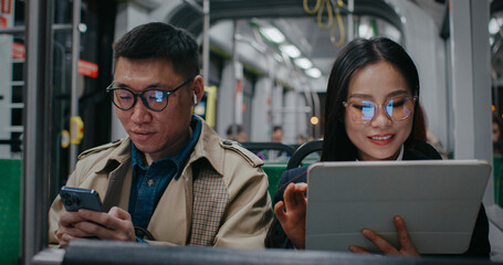 Cute Asian couple traveling together after work. Sitting together but using separate devices. Man busy typing on smartphone while girl playing games or scrolling websites on Internet on tablet device.