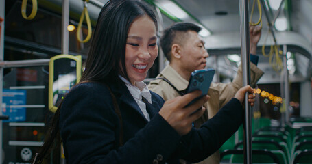 Asian girl trying to keep balance in moving bus or train. Holding with one hand by metal tube. Starting to laugh from video on her smartphone. Man besides beginning to laugh after taking peek.