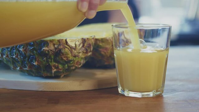 Close up of woman's hand pouring fresh pineapple juice into a glass with a pineapple sliced in half in the background in a brightly lit kitchen. Studio shot. High quality 4k footage