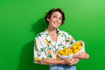 Photo of beautiful young dreamy girlfriend holding handmade basket for organic fresh fruits lemon citrus isolated on green color background - obrazy, fototapety, plakaty