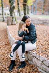 Mom hugs a little girl with an apple in her hands sitting on her lap on a low fence in the park - obrazy, fototapety, plakaty