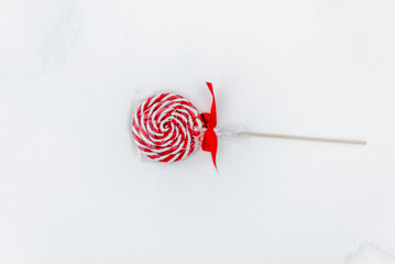 A festive and wintery scene  of a red and white round flat lollipop lying on the snow, surrounded...