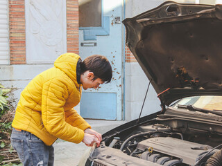 A young caucasian guy in a yellow jacket inspects an internal breakdown with an open car hood - obrazy, fototapety, plakaty