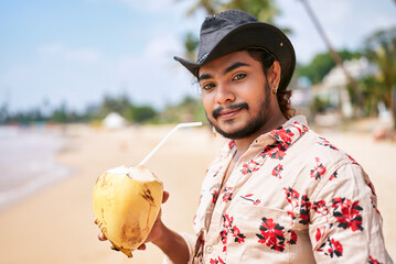 Smiling gay man in floral shirt and cowboy hat enjoys fresh coconut drink on sunny tropical beach....