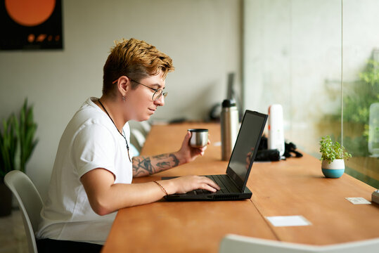 Transgender employee in modern office sips coffee from metal mug, works focused on laptop. Inclusive workplace scene with tattooed pro, potted plant, bright workspace. - Powered by Adobe