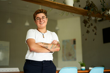 Confident Gen Z individual with tattoos stands in modern office. Transgender young adult smiles,...