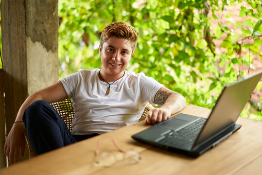 Transgender Gen Z with tattoos smiles, works on laptop in tropical coworking space. Inclusive remote work scene captures gender diversity, modern freelancing trend amongst rich plants.