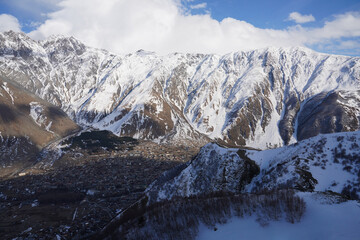 Snow-capped mountain range peaks under blue sky and white clouds  with a village below, sunny day, Gergeti Trinity Church, Stepantsminda, Georgia