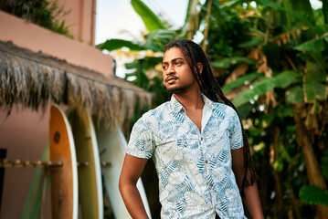 Dark-skinned surfing coach standing by thatched hut, surfboards lined up. Calm, confident...