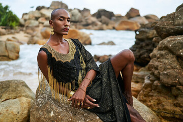 Non-binary black person in luxury dress on rocks in ocean. Trans ethnic fashion model wearing jewellery dressed in posh gown poses gracefully in tropical seaside location portrait. Pride month.