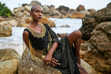Non-binary black person in luxury dress on rocks in ocean. Trans ethnic fashion model wearing...