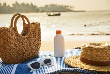 Beach essentials laid on a striped towel by the sea at sunset include a straw hat, sunglasses, sun...