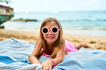 Smiling girl with pink sunglasses lies on blue striped towel at sunny beach. Happy child enjoys...
