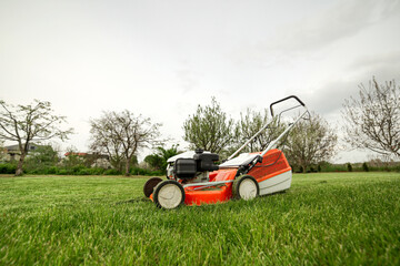 side view of modern orange-grey gasoline lawn mower cutting bright lush green grass. Gardening work tools. Rotary lawn mower machine on lawn. Professional lawn care service. Place for text