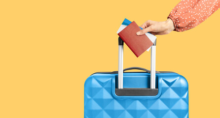 female hand holding the handle of a suitcase and holding a passport and tickets on a yellow...