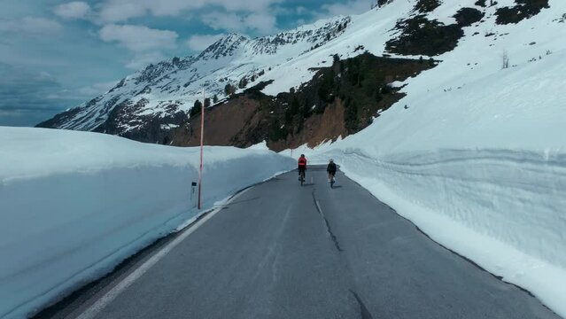 Two cyclists pass camera during descend on mountain road. Road cleared of snow in spring, creating snow corridor walls on the sides. Cinematic cycling in winter
