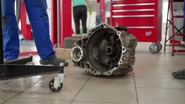 Auto mechanics in uniform remove car gearbox at repair shop. Teamwork in vehicle maintenance, manual transmission system servicing. Hands-on training in automotive tech, precise machinery work.
