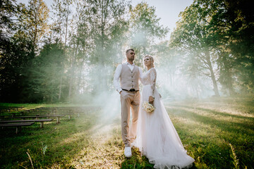 Valmiera, Latvia - August 10, 2023 - Bride and groom standing in a sunlit forest clearing with...