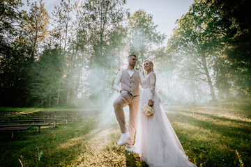 Valmiera, Latvia - August 10, 2023 - Bride and groom stand amid misty forest clearing, sunbeams...