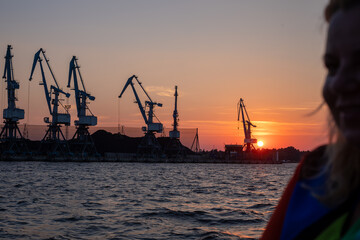 Sunset at the harbor, cranes silhouetted against the evening glow, with a womans profile in the...