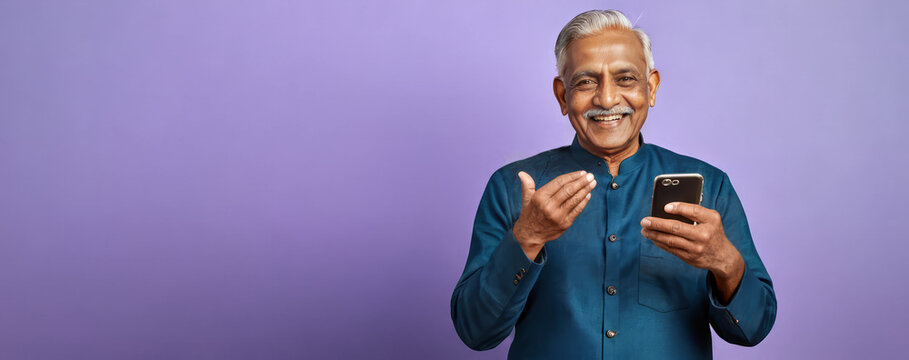 Panoramic wide banner of senior Indian man with blue shirt, grey hair and moustache smiling and holding mobile phone isolated on purple studio background, copy space for text - Powered by Adobe