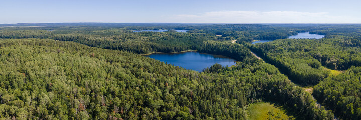 Panoramic aerial view of a vast boreal forest with small blue lakes under a pale blue sky. A road runs through the forest.
