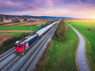 Aerial view of freight train moving near alpine village, mountains and green fields, bike path at...