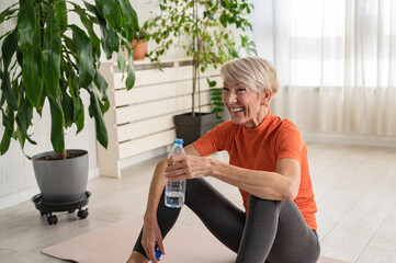 Beautiful senior woman drinking water after exercise. Active senior woman practicing yoga indoors....