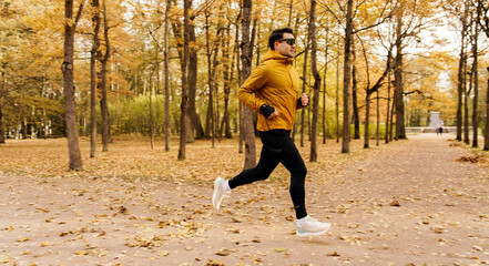 A man in a yellow jacket and sunglasses runs energetically through an autumnal park with fallen...