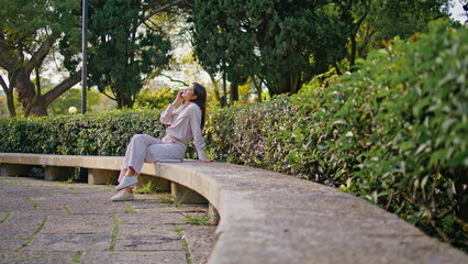Cheerful woman enjoy phone conversation sitting on park bench at green foliage