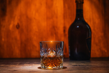 A glass of whiskey in the foreground of a scene with a wine cellar, a bottle of alcohol and an oak...