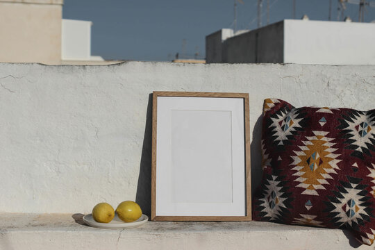 Blank vertical wooden frame picture mock up. Fresh lemon fruit, Mexican pattern cushion. White old textured white wall in sunlight. Front view, no people. Summer display background for art, posters.