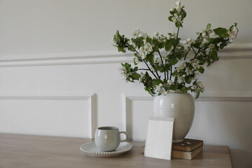 Springtime breakfast still life. Empty greeting card, invitation mockup. White ceramic vase with blooming apple tree branches. Cup of coffee, tea on wooden table, old books. Scandi home interior
