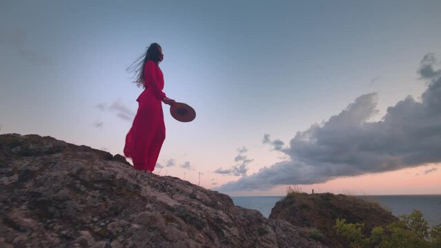 Amazing view, a young woman in a red dress standing on a rock against the backdrop of the sea or ocean and sunset. An attractive girl in nature against the backdrop of a landscape during the wind