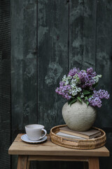 Moody farm spring breakfast still life. Purple, white lilac flowers bouquet in textured vase with cup of coffee, tea. Wicker tray, vintage books. Blurred old green wooden door background. Vertical.